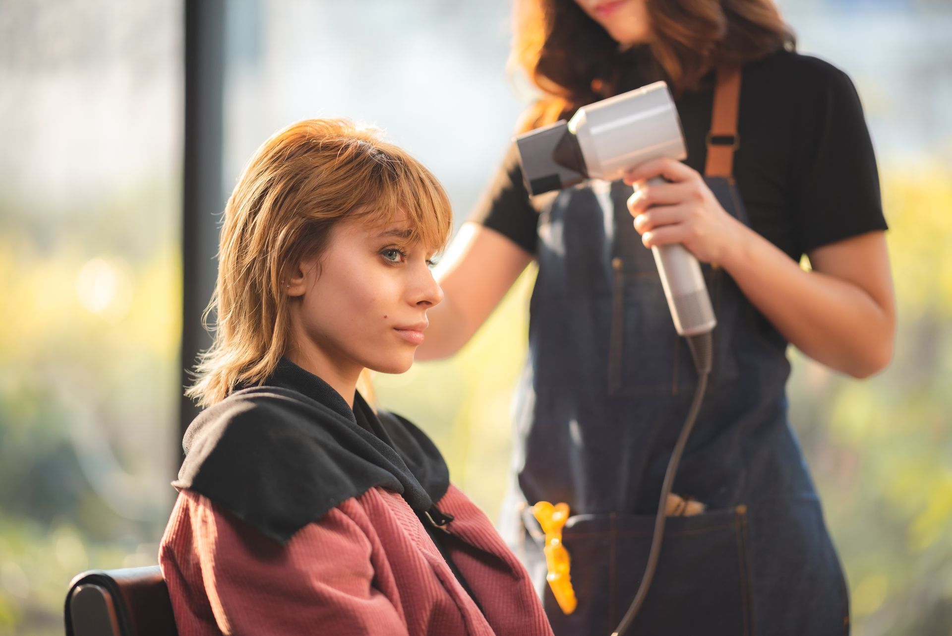Woman having hair blow-dried by stylist in salon; sunlight streams through a window.