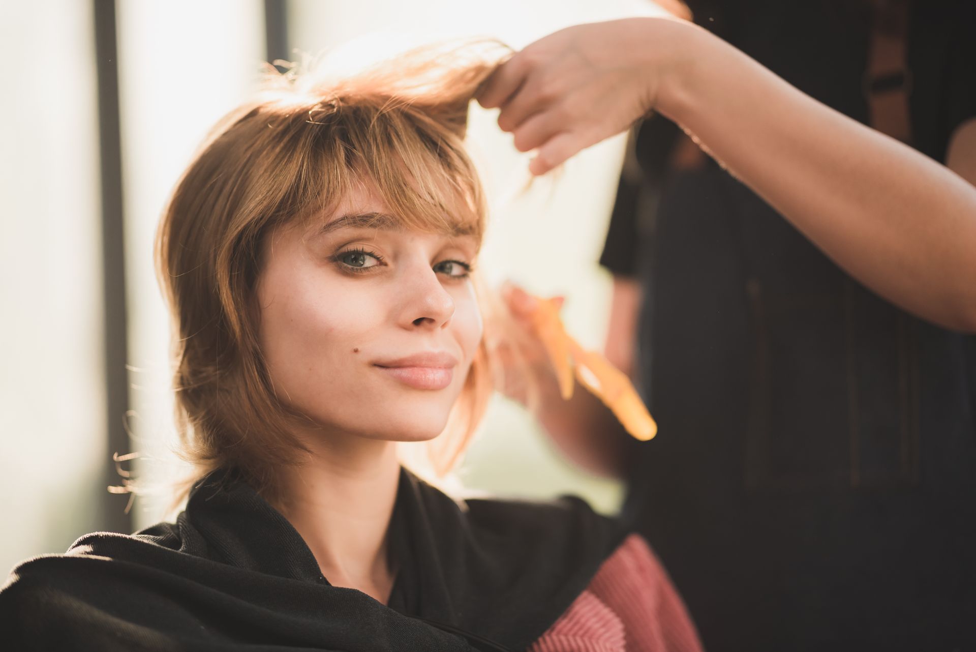Woman getting a haircut, smiling at camera, with hands holding her hair in a salon.