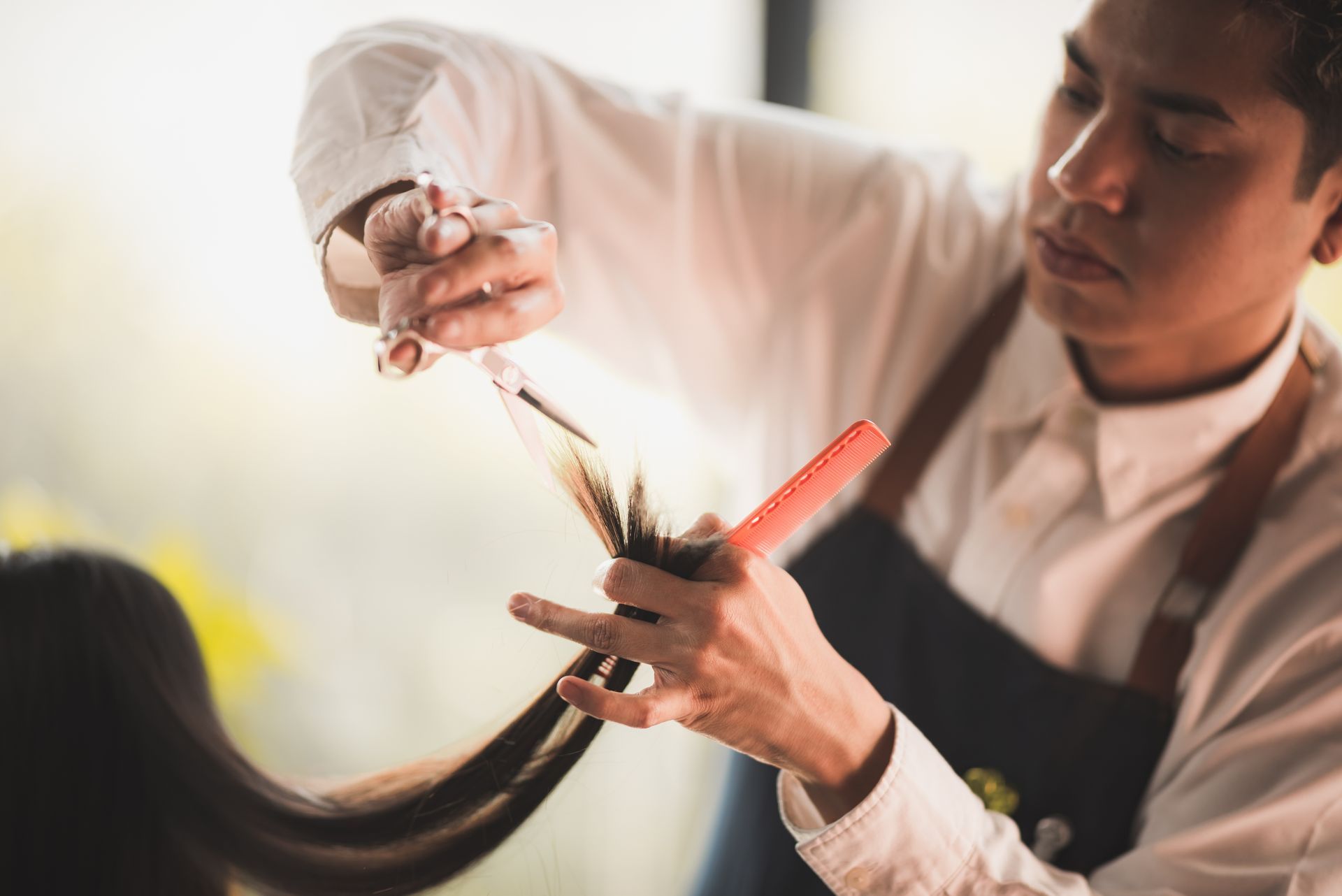 Barber cutting a client's hair with scissors and comb. Indoors, focus on hands, white shirt, and apron.