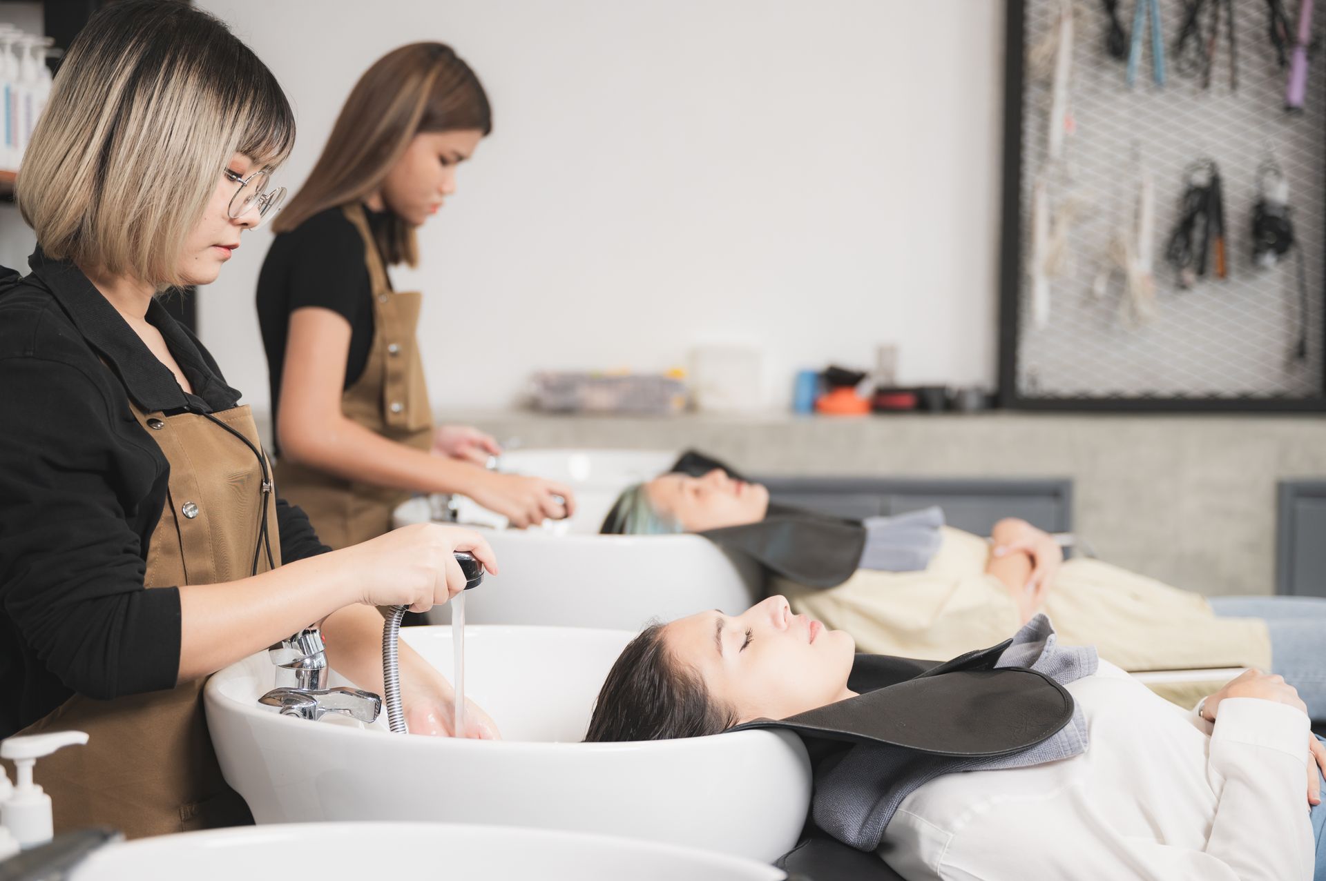 Hair stylists washing clients' hair in a salon. Clients recline in bowls, while stylists attend to them.