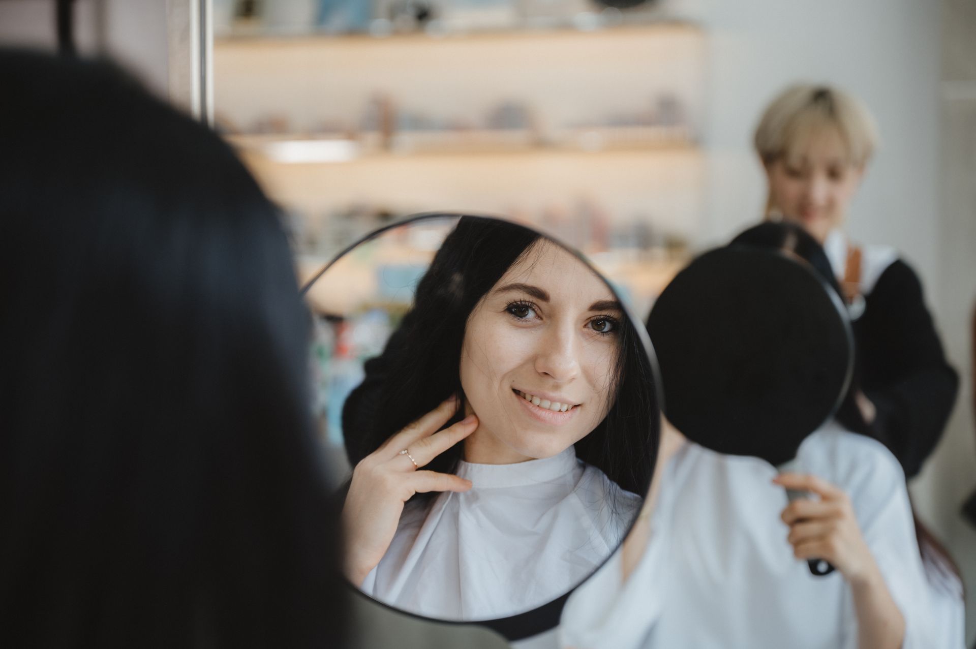 Woman smiles at her reflection in a mirror held by a stylist in a salon.