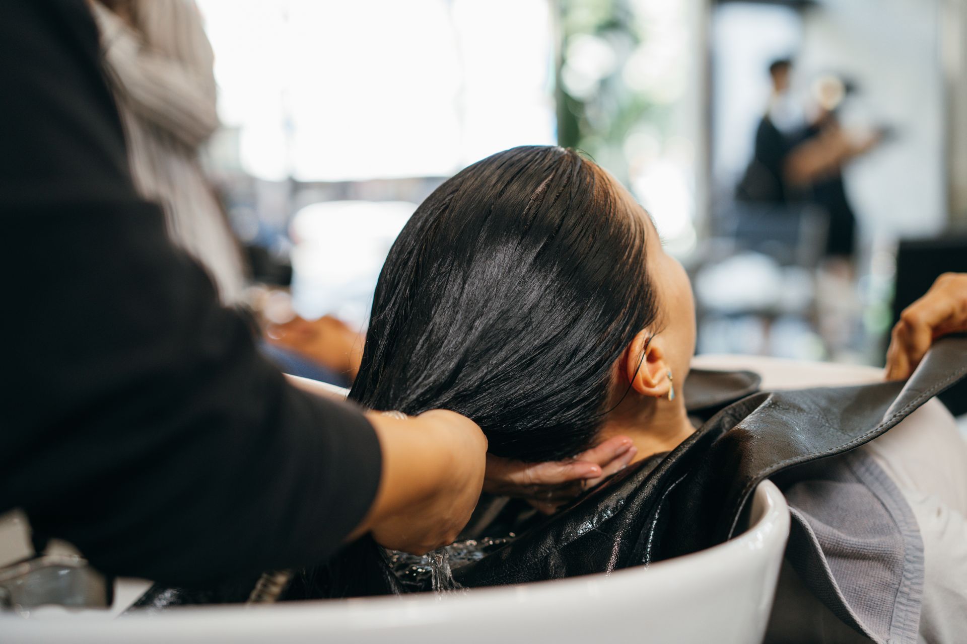 Woman having her hair washed at a salon; stylist's hands working, white sink, black cape.