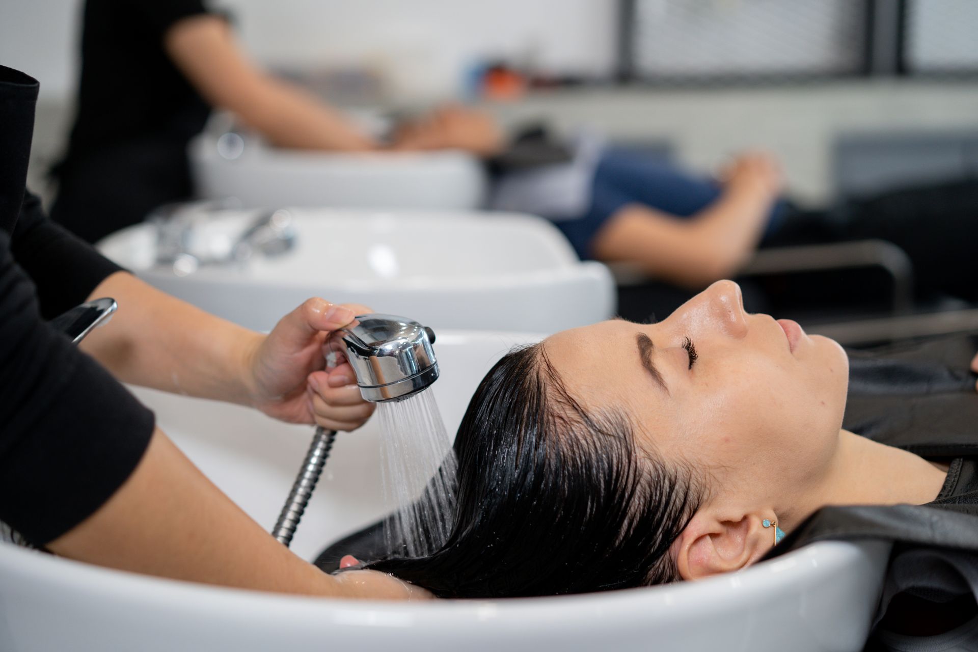 Woman having hair washed at a salon; stylist's hands holding sprayer, water flowing onto her dark hair.