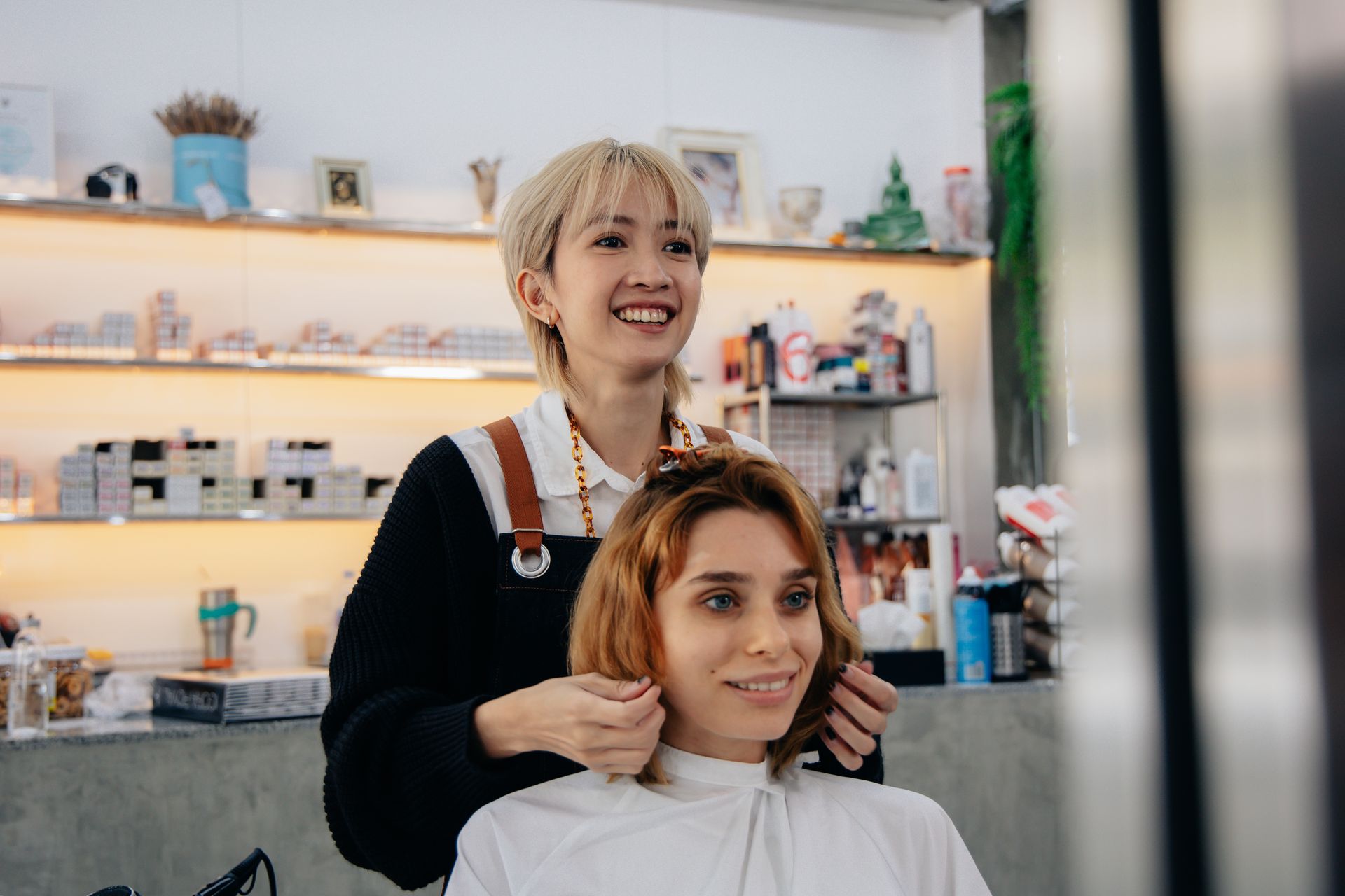 Hair stylist smiling, arranging client's hair in a salon. Blonde hair, black apron, white cape.