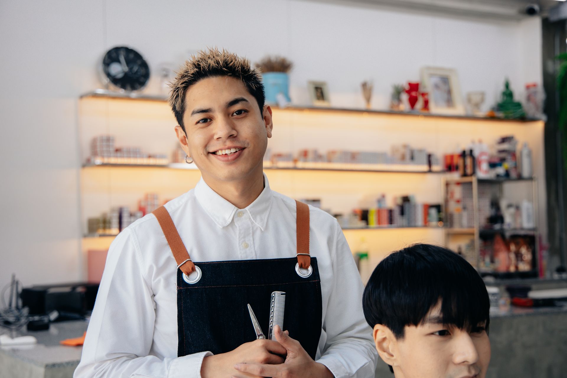 Hairdresser with blonde-streaked hair smiles while holding shears, cutting a client's hair in a salon.