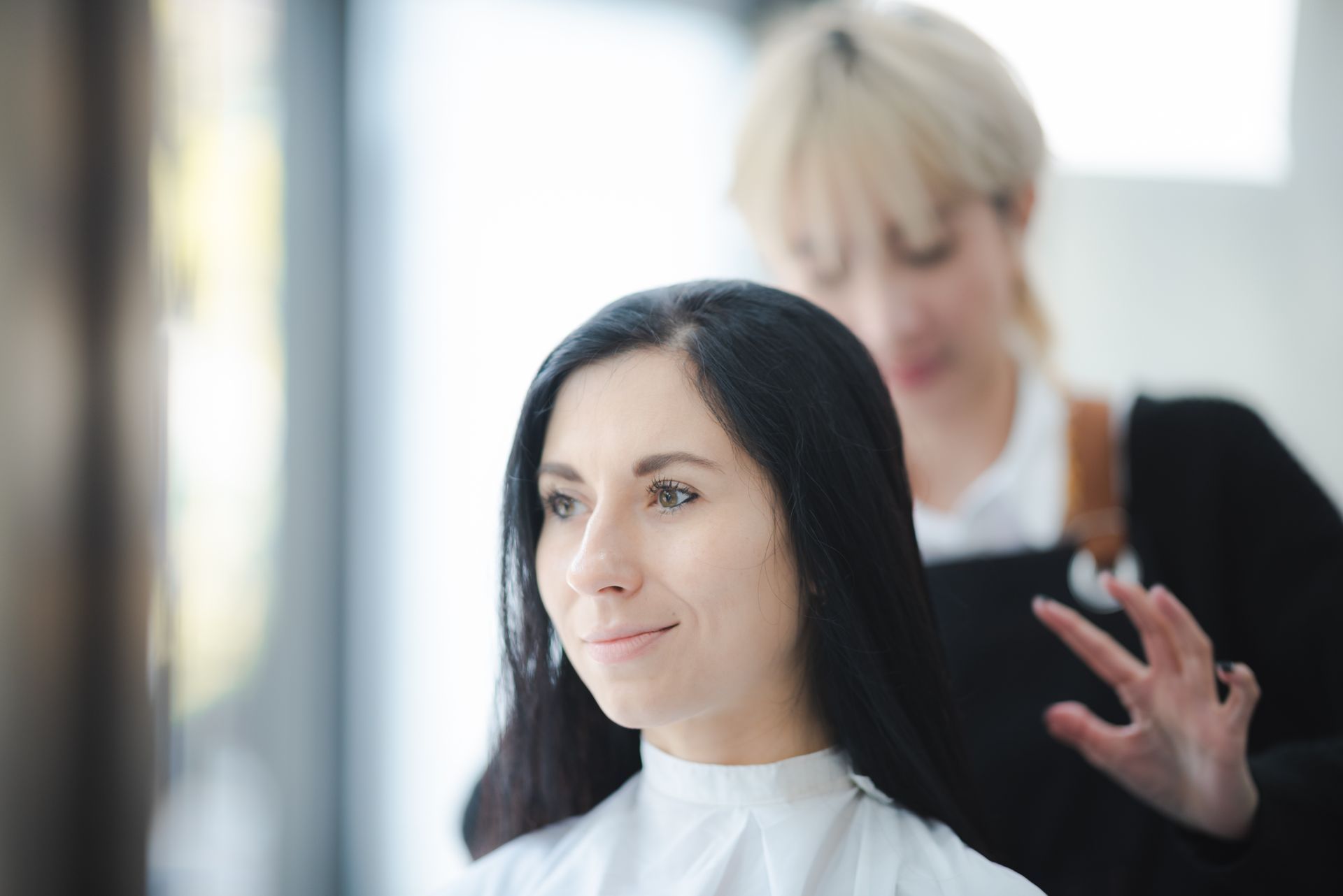 Woman with black hair getting it styled by a blonde hairdresser in a salon.