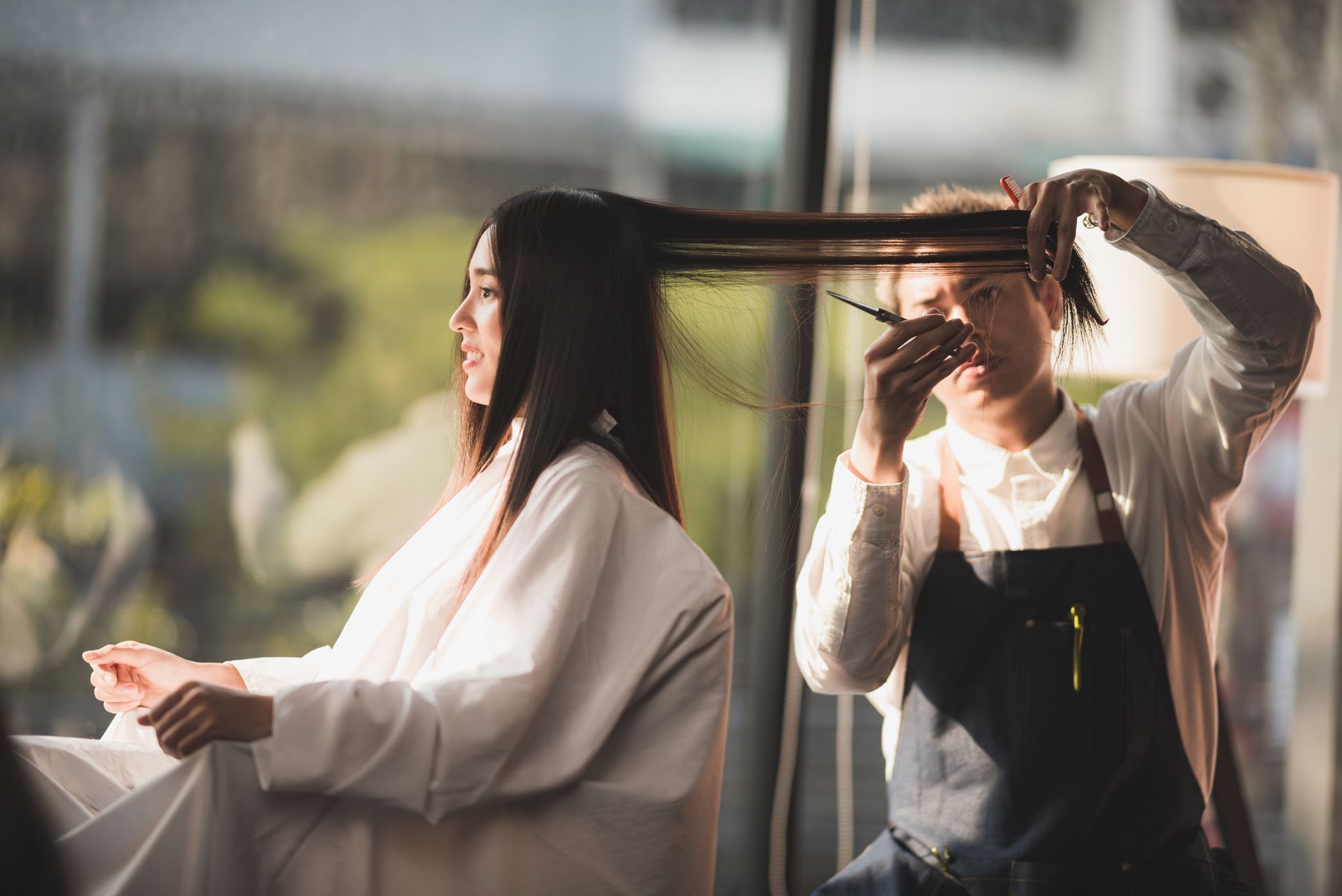 Woman getting haircut at salon; stylist cutting long brown hair near window.