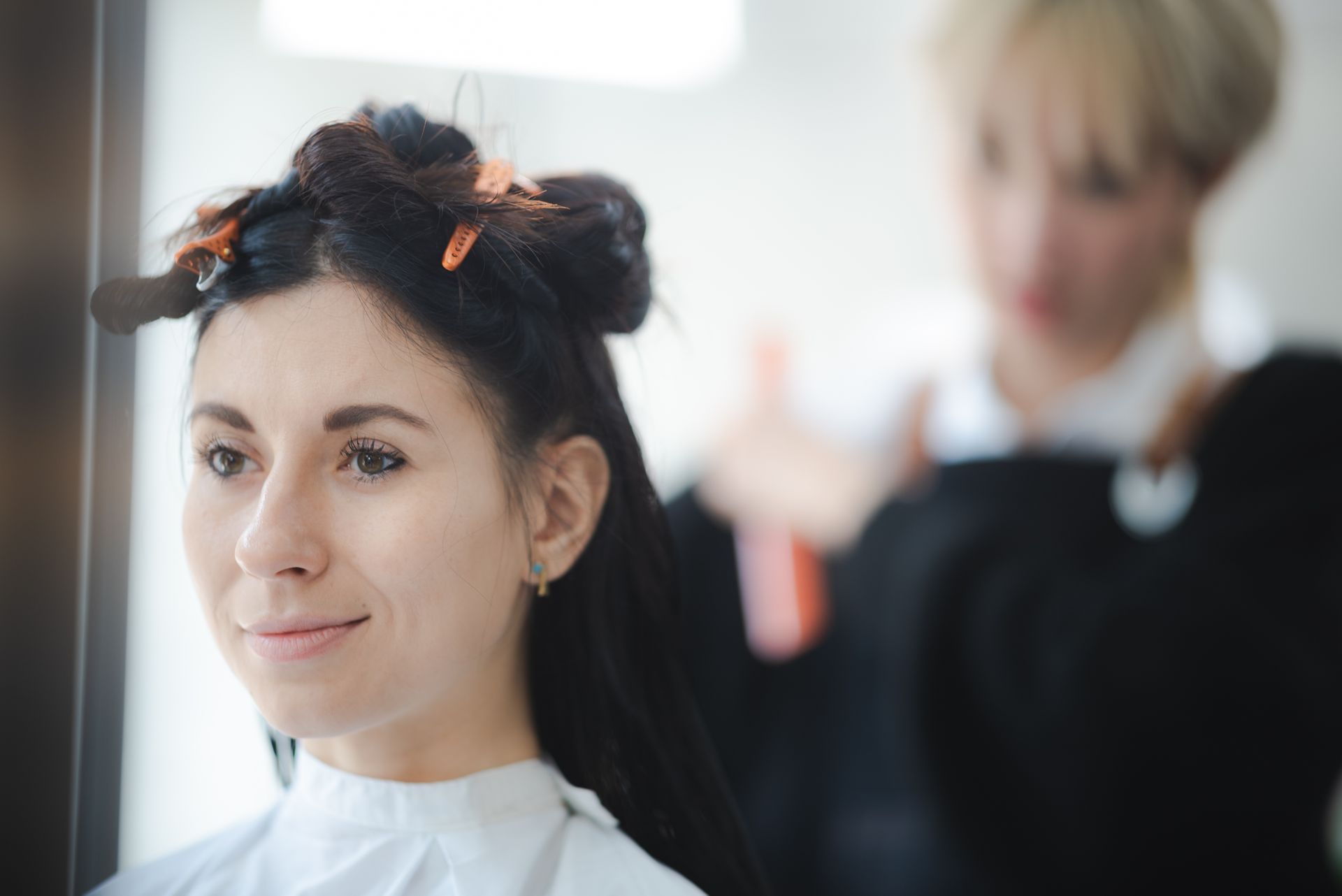 Woman with dark hair in hair rollers at salon; stylist in background.