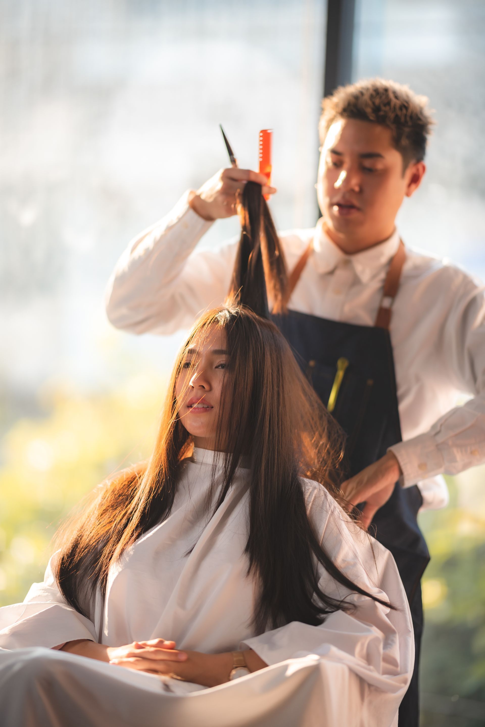 Hairdresser cutting a woman's long, dark hair in a salon; sunny setting, white cape and shirt, blue apron.
