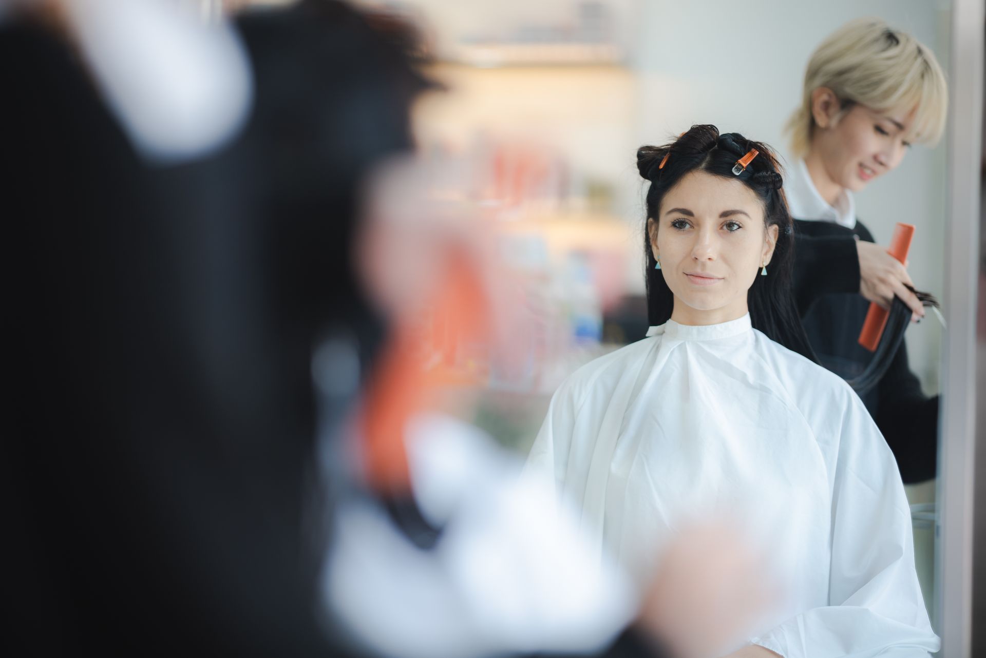 Woman in white cape having hair styled at salon. Stylist in background.