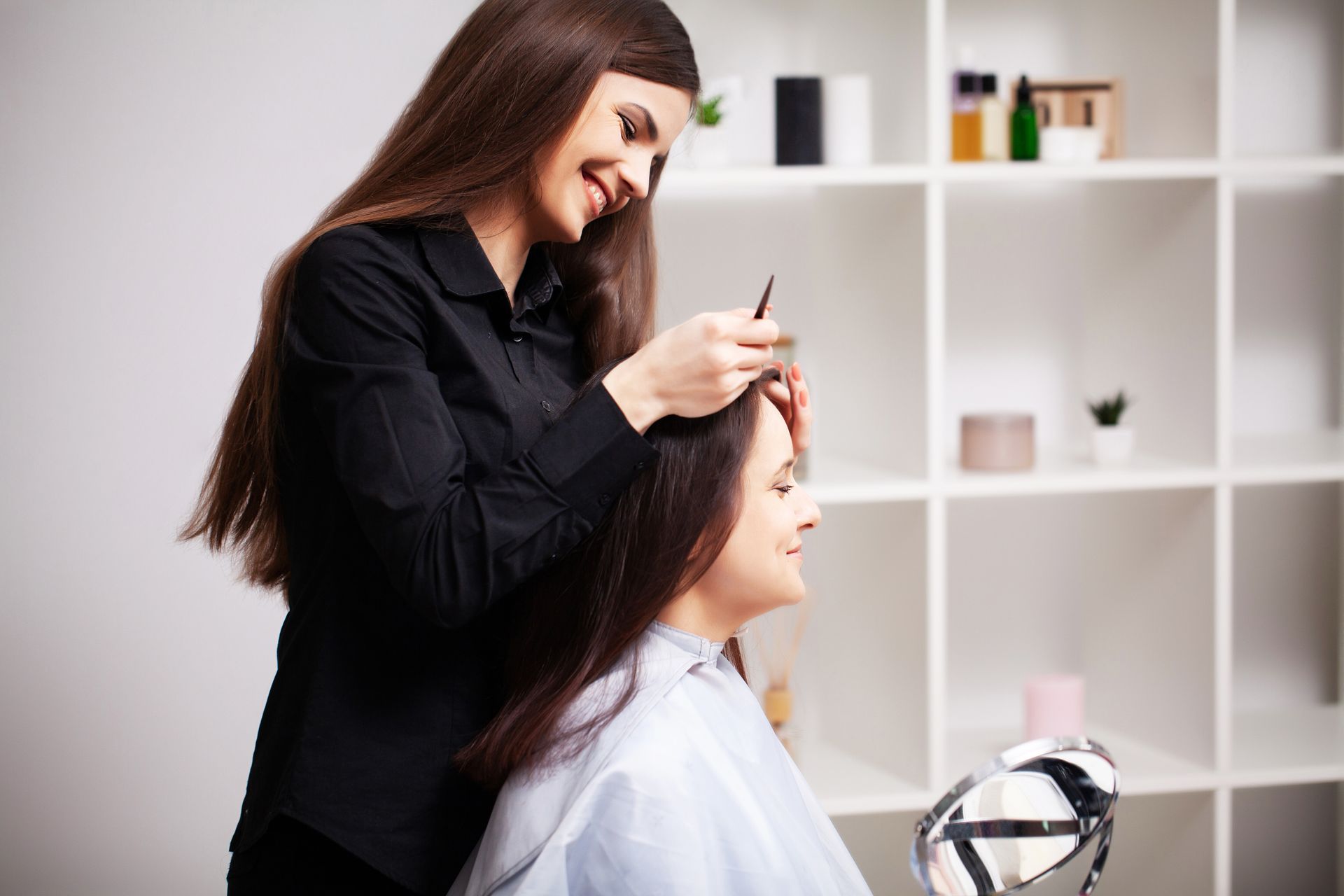 Woman getting her hair styled in a salon; stylist combs client's hair. Neutral background with shelving.