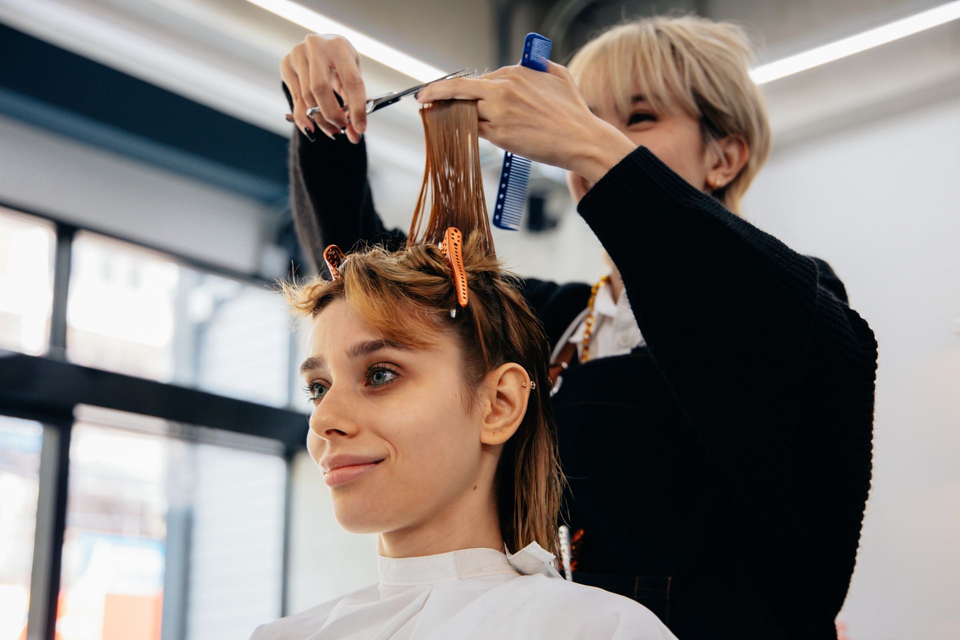Hairdresser cutting a woman's hair, indoors. The woman smiles, wearing a cape. The hairdresser holds scissors.