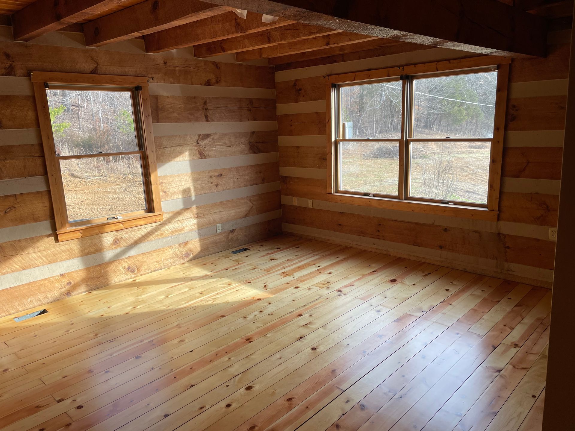 An empty room in a log cabin with wooden floors and two windows.
