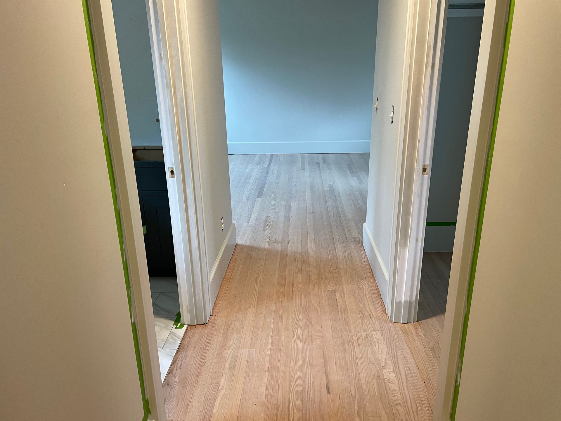 A hallway with hardwood floors and two doors leading to a room.