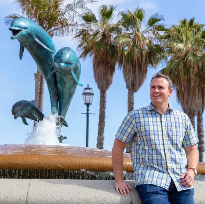 Stephen leans against a wall in front of a fountain with dolphins on it