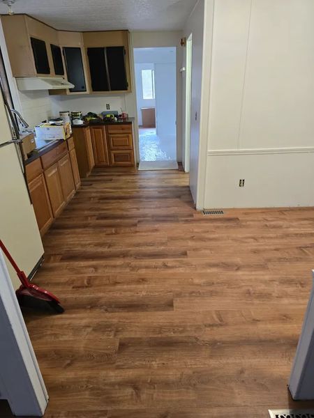 Kitchen with wood-look flooring and light brown cabinets. A broom leans against the refrigerator. A doorway leads to a hallway and other rooms.