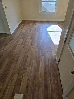 A room with wood-look flooring, cream walls, and a window letting in sunlight. A white door is on the right.