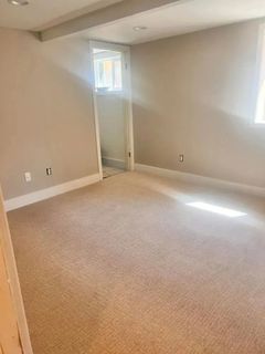 Empty beige-walled room with beige carpet, a small window, and a doorway to another space. Sunlight streams in from a window.