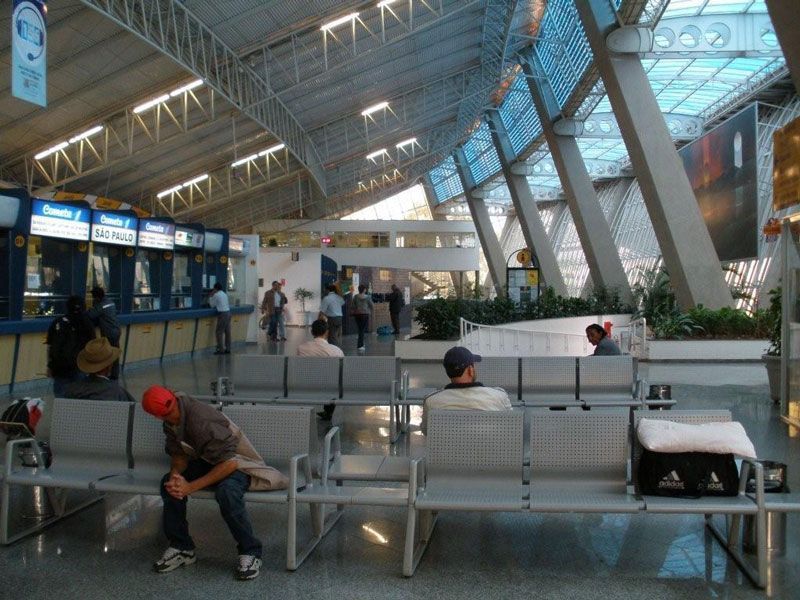 Táxis brancos enfileirados sob a cobertura de um prédio; plantas verdes e um céu azul se destacam.