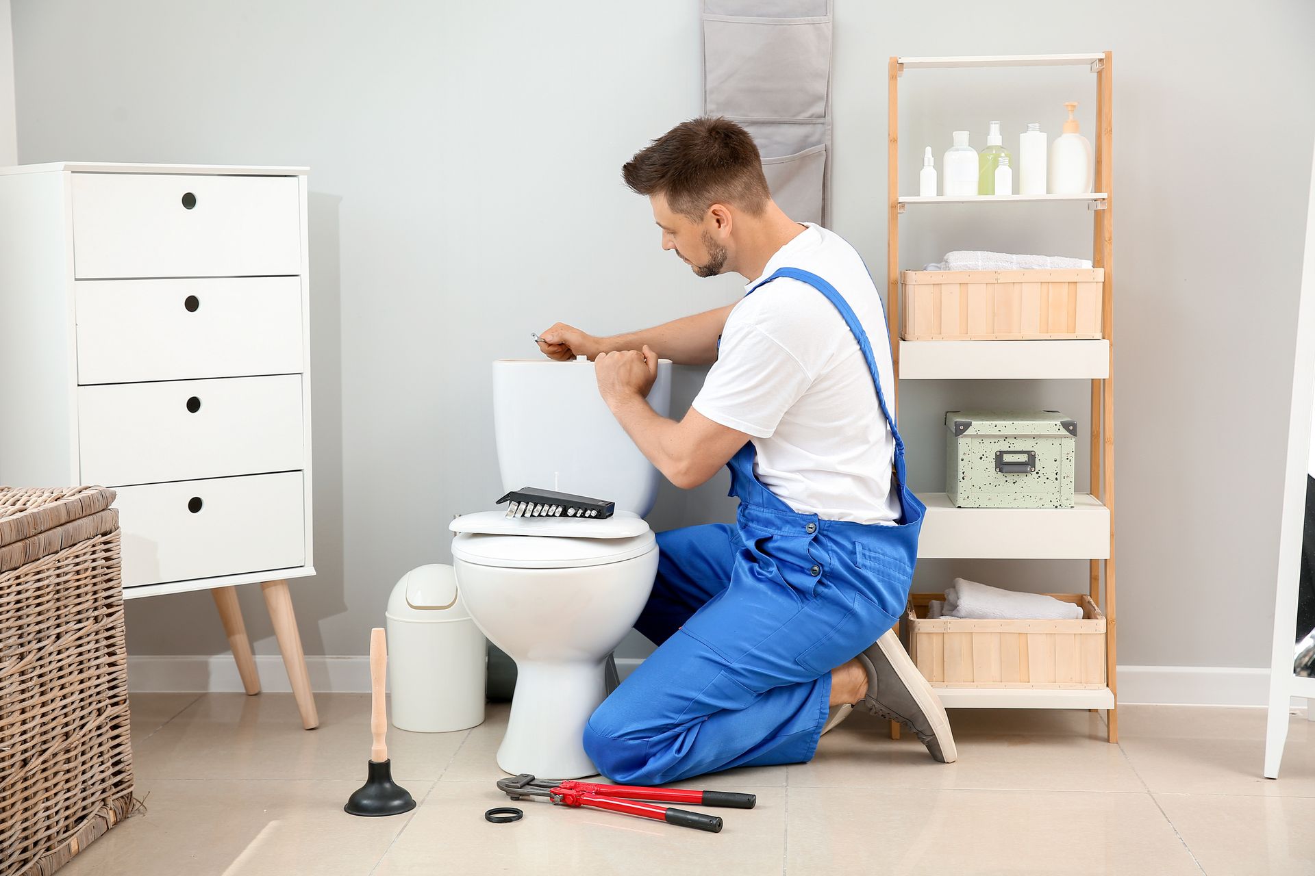 Plumber kneeling beside a toilet, using tools in a bright bathroom.