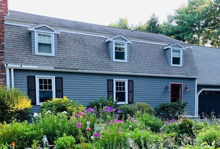 A blue house with a gray roof and black shutters is surrounded by flowers and bushes.