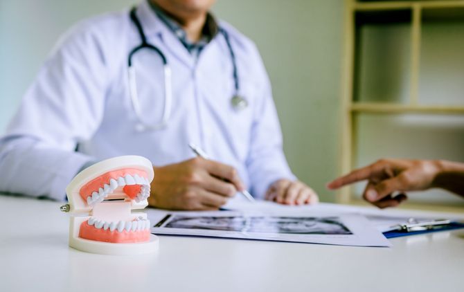 Dentist in white coat discusses x-rays with a patient, pointing to the image, alongside a model of teeth.