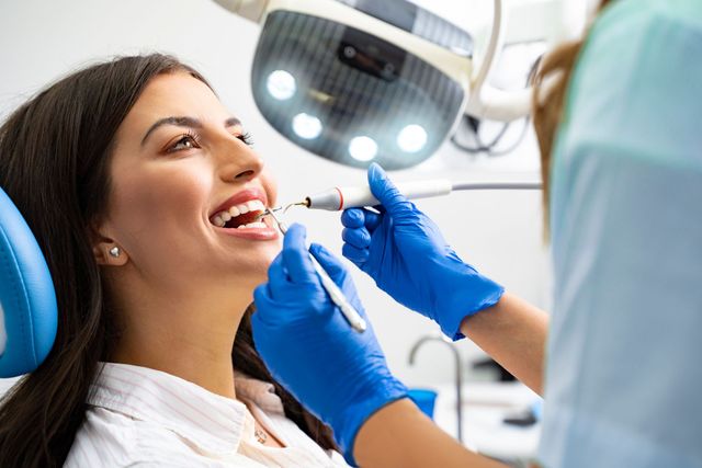 Woman at the dentist, having teeth cleaned. Dentist in blue gloves holds tools. Bright lighting and equipment.