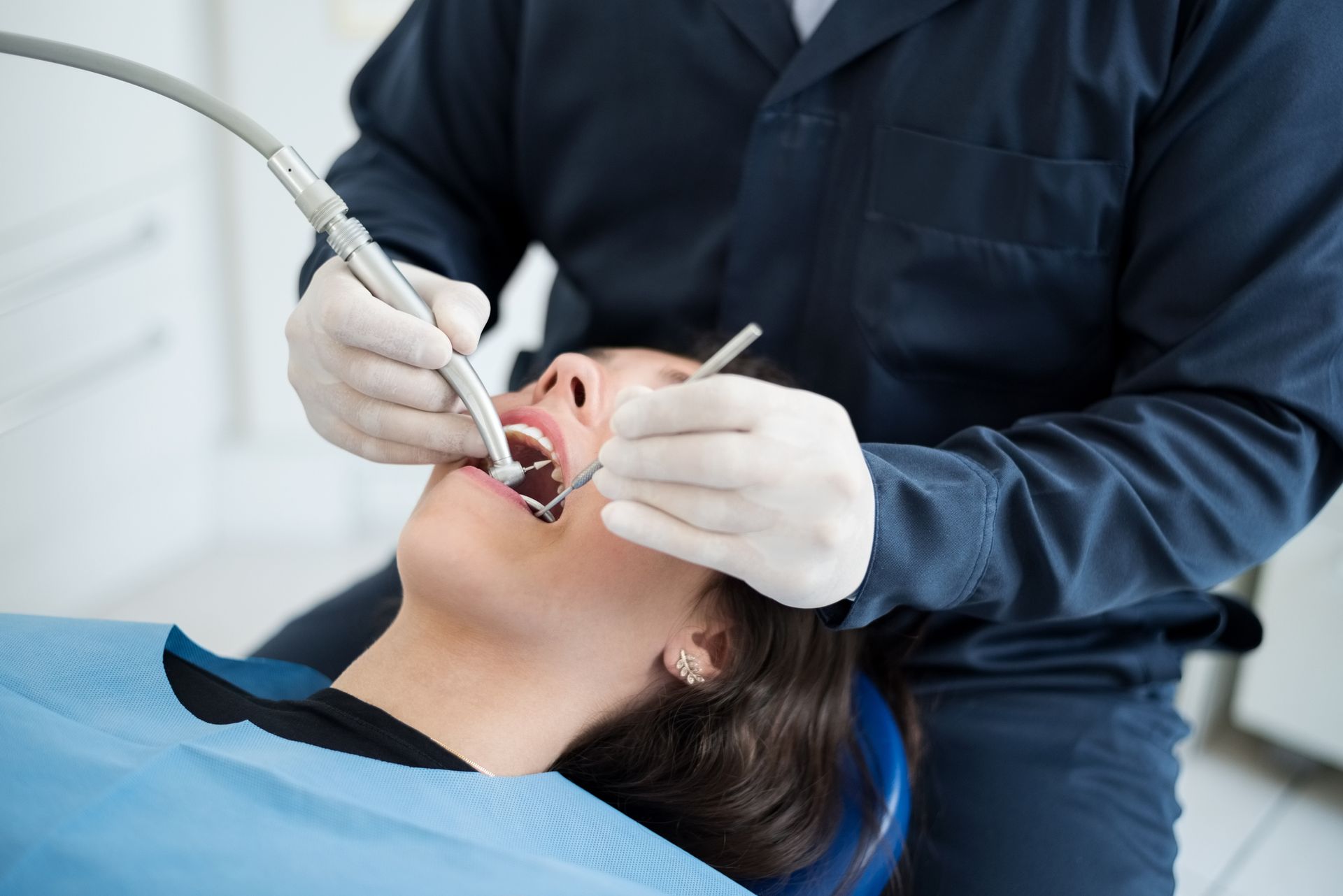 Dentist examining a patient's teeth. The patient reclines in a blue chair, mouth open. Dentist wears gloves and a dark blue outfit.