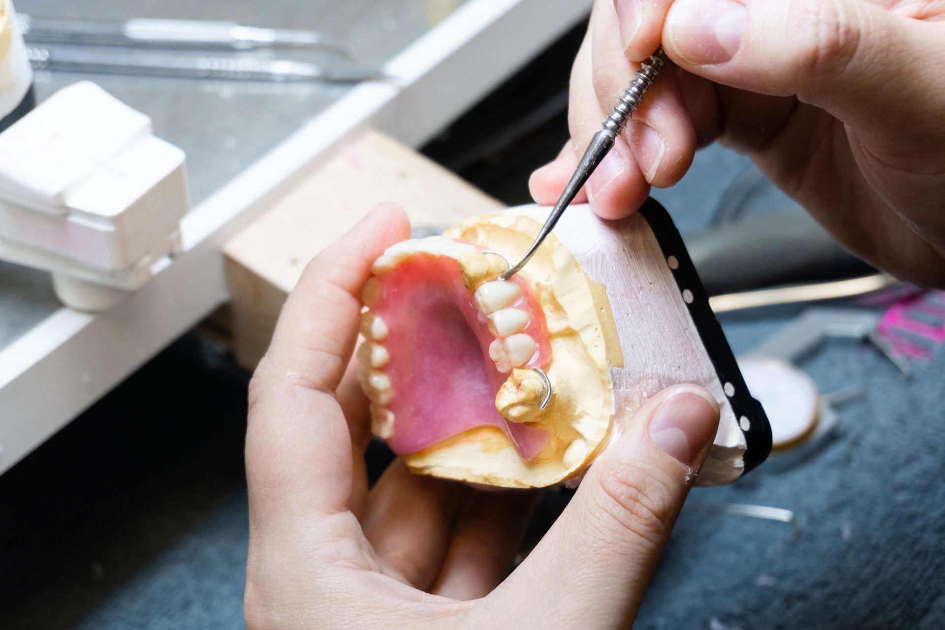 Hands working on a dental mold with teeth and pink material, using a small tool.