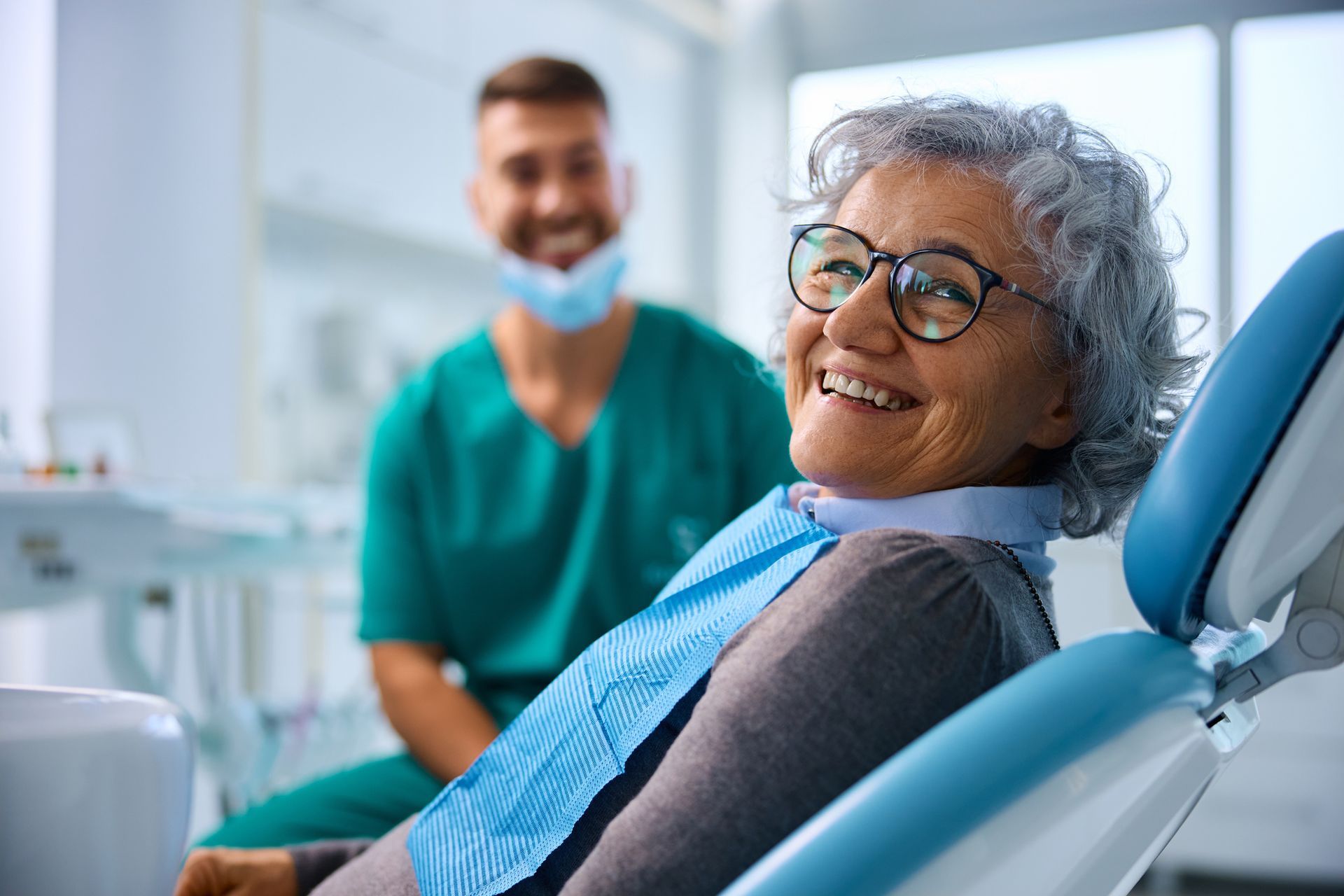 Elderly woman smiles in dentist chair, blue bib, doctor in green scrubs smiles in background.
