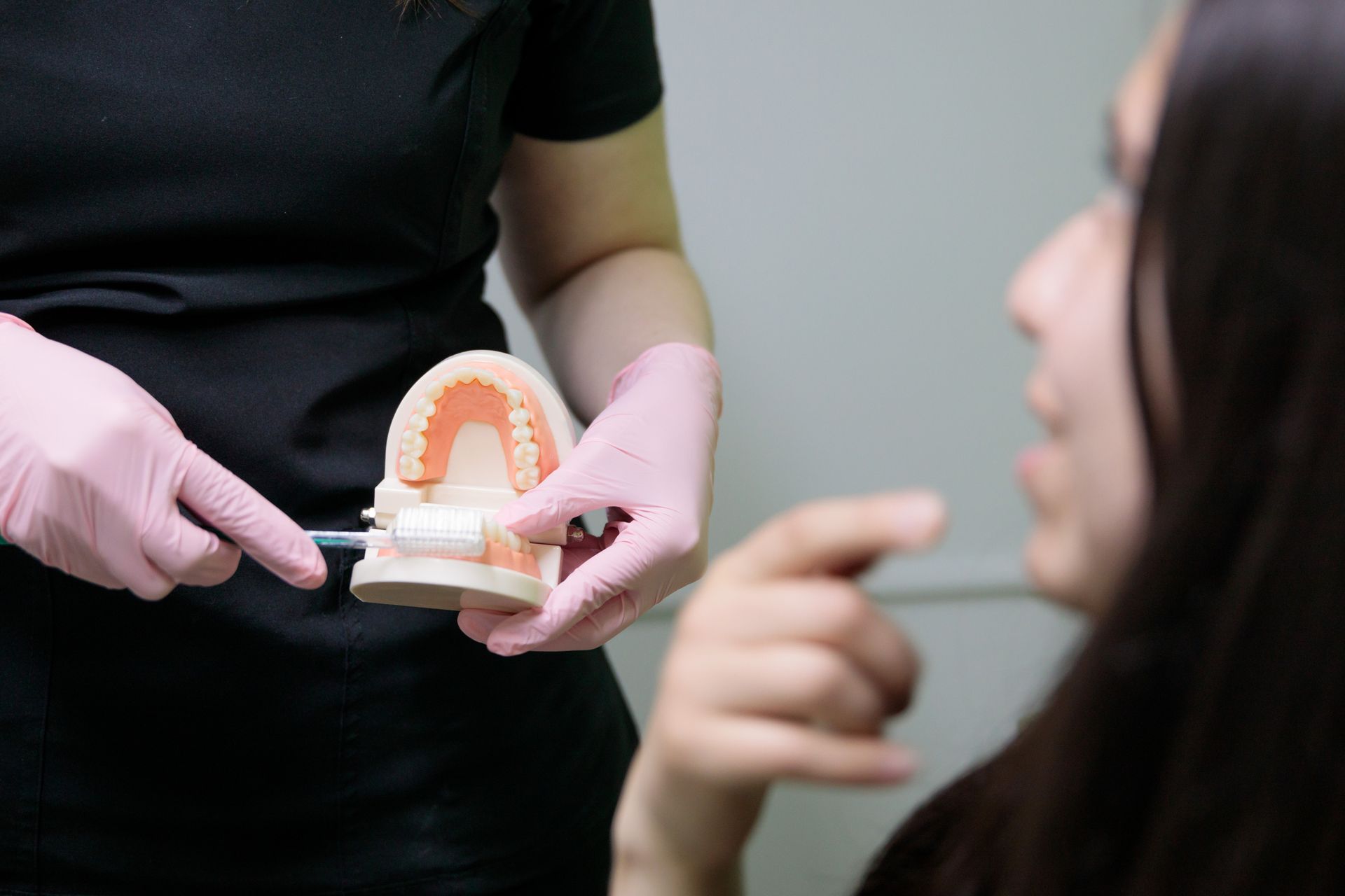 Dentist demonstrating brushing technique on teeth model to a patient. Pink gloves, black shirt, white background.
