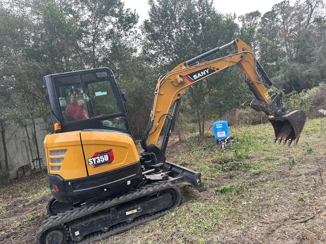 A yellow Sany mini excavator sits on a grassy field with an operator inside, positioned near trees and a blue item.