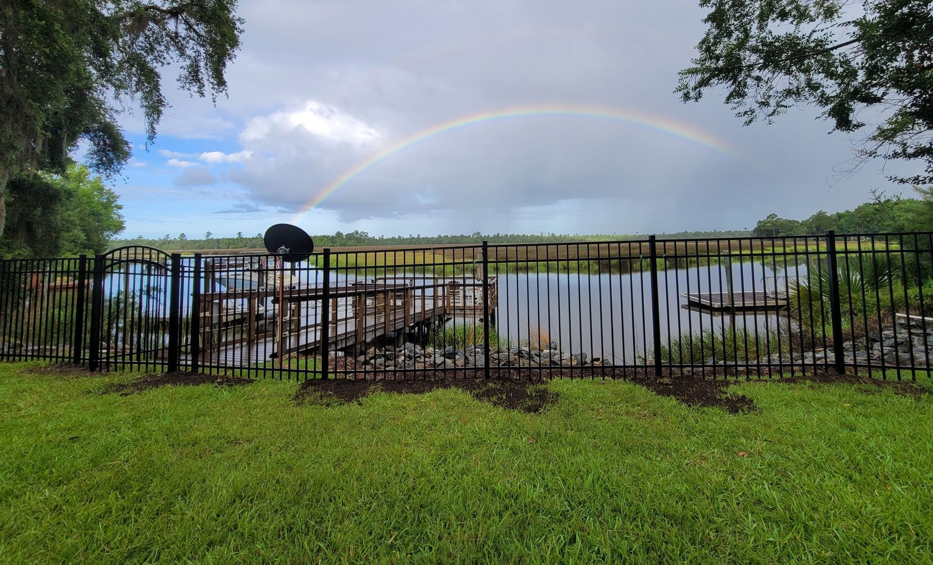 A rainbow arches over a pond seen through a black metal fence, with green grass in the foreground and a cloudy sky.