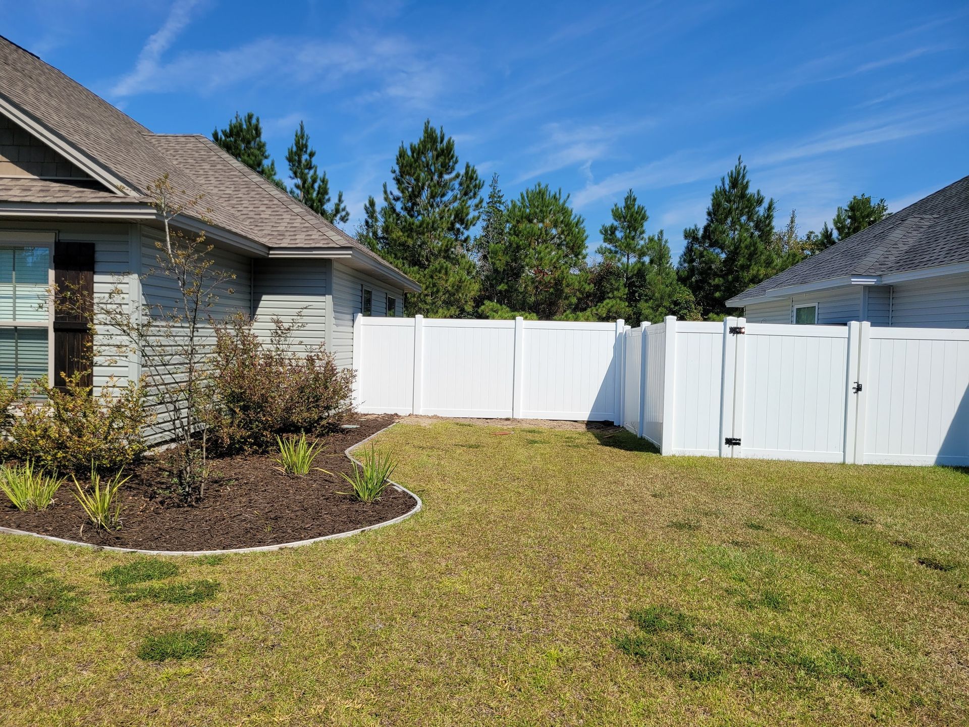 A residential backyard with a gray house, landscaped mulch bed, and a white privacy fence against a blue sky.
