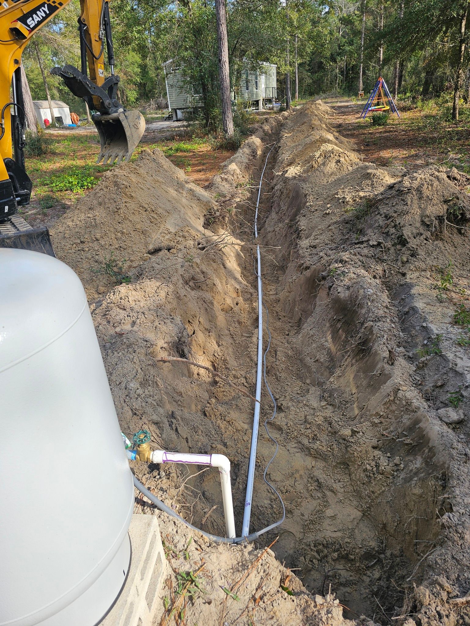 A backhoe digs a trench for a white pipe connected to a large, grey water storage tank in a wooded, outdoor area.