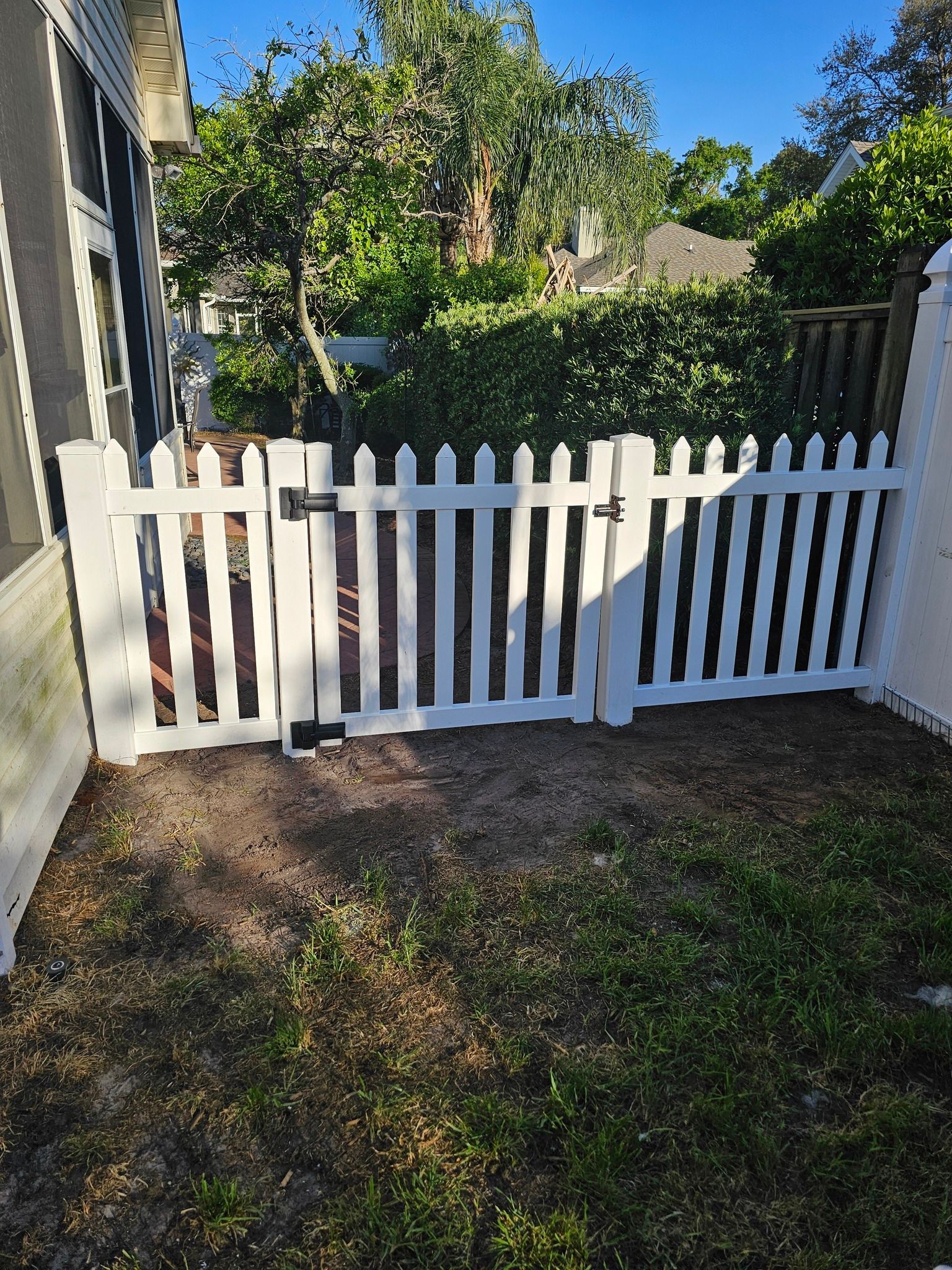 A white vinyl picket fence with a gate, located outdoors in front of a house with a screened porch and green foliage.