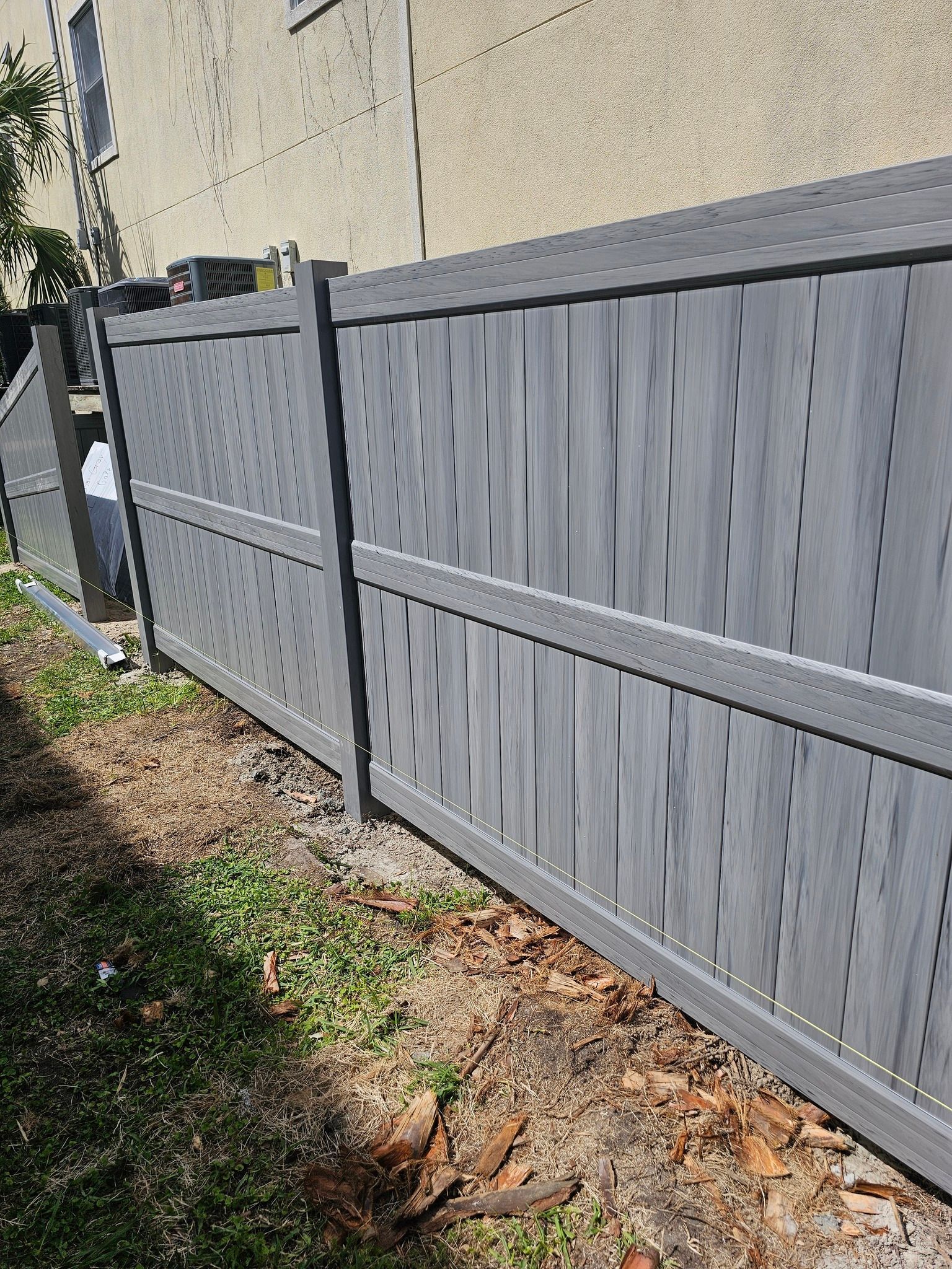 A grey vinyl privacy fence running along the side of a building, over a dirt ground with sparse grass.