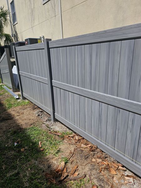 A gray, vertical-slat privacy fence stands alongside a beige building wall with dry leaves and grass on the ground.