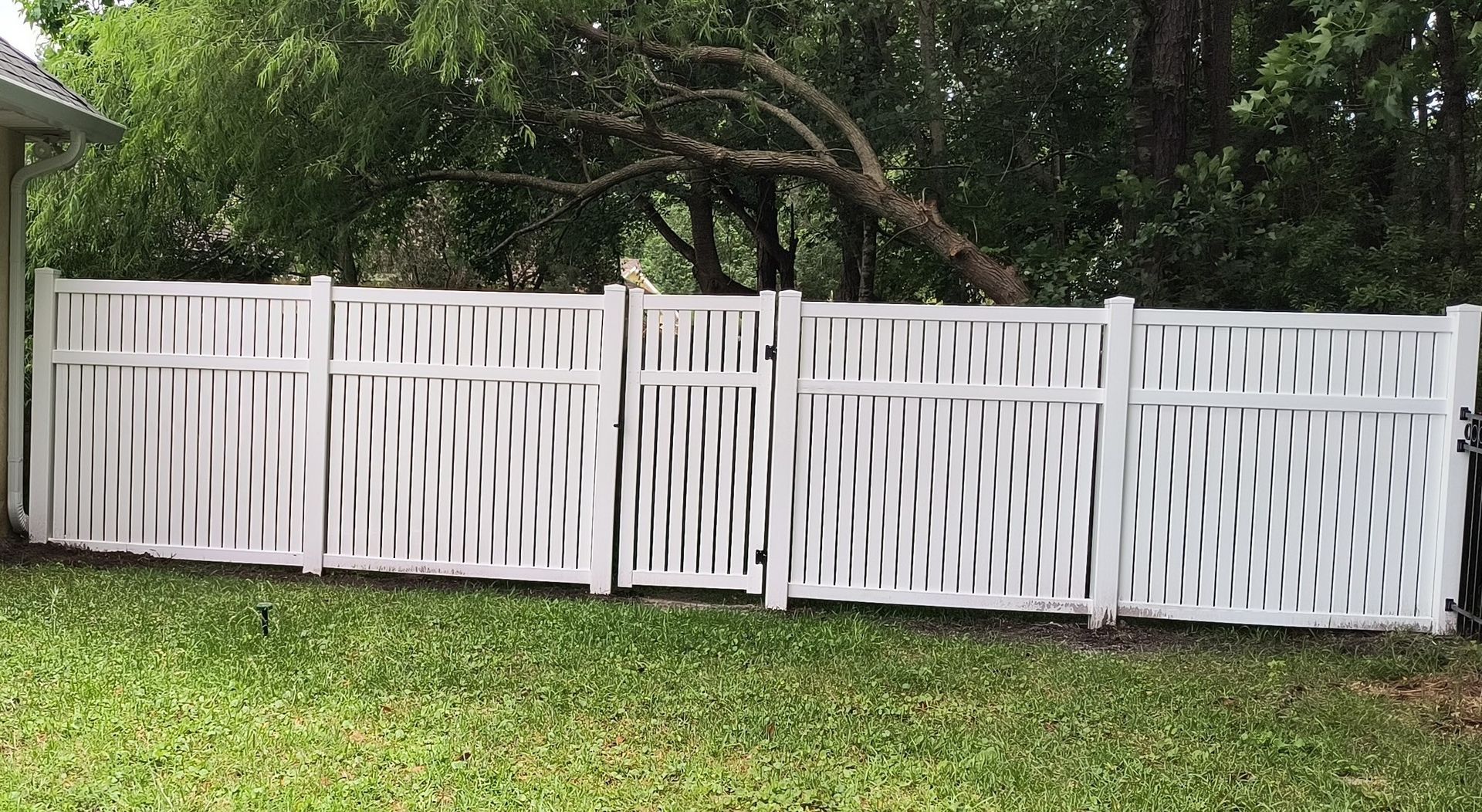A white vinyl fence with a basket-weave pattern and a centered gate, situated on a lawn in front of green trees.