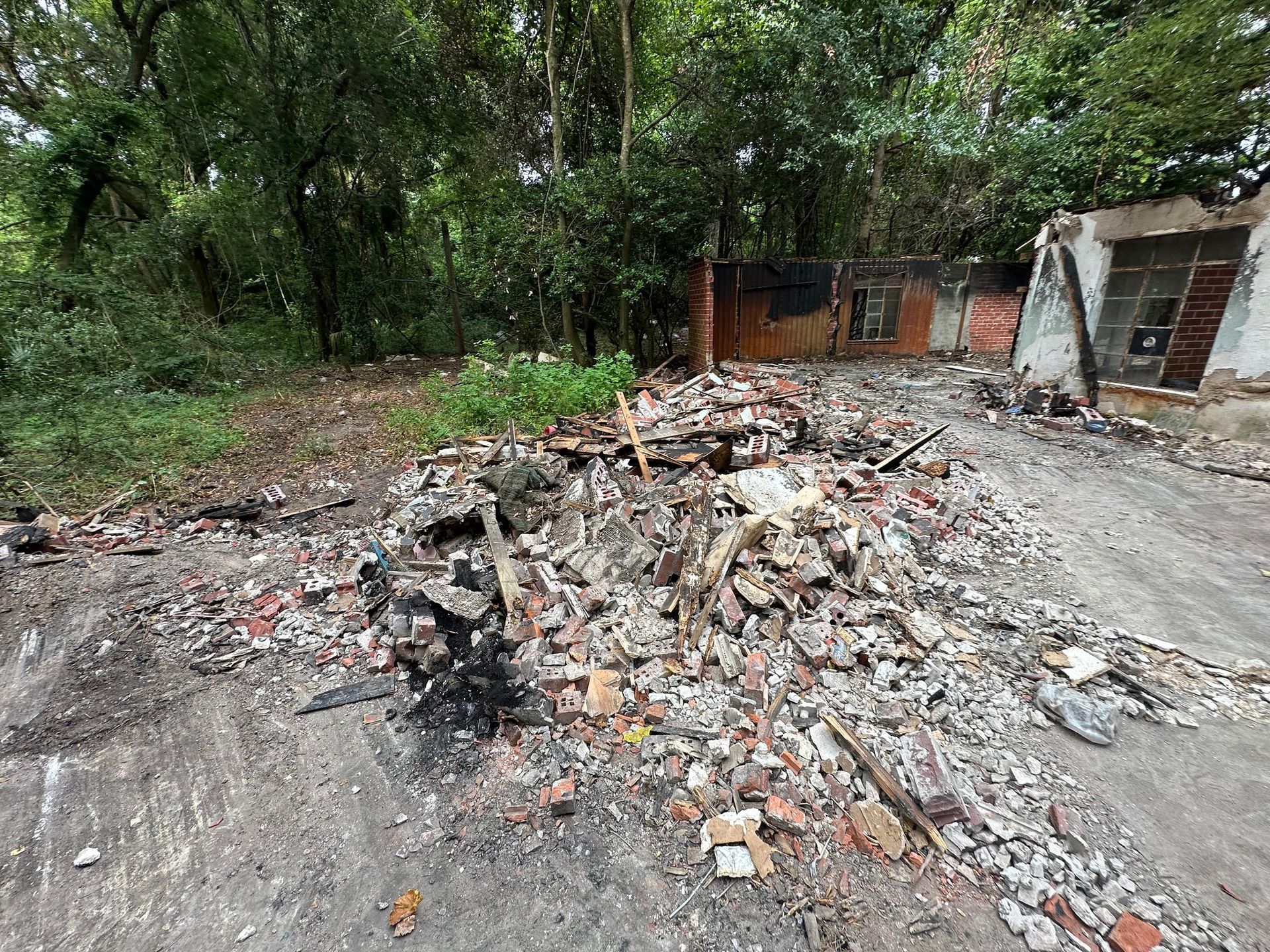 A pile of debris and rubble in front of the partial remains of a building, with a dense forest in the background.