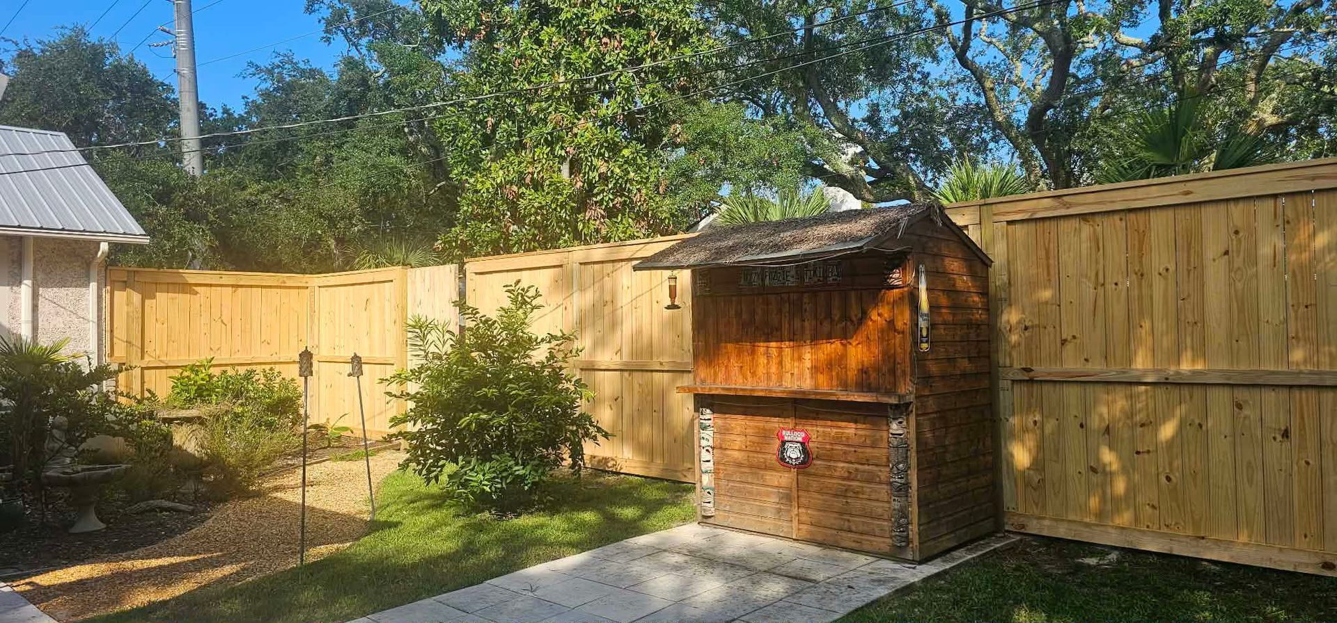 A small, wood-stained tiki bar structure stands in a backyard corner against a tall wooden privacy fence.