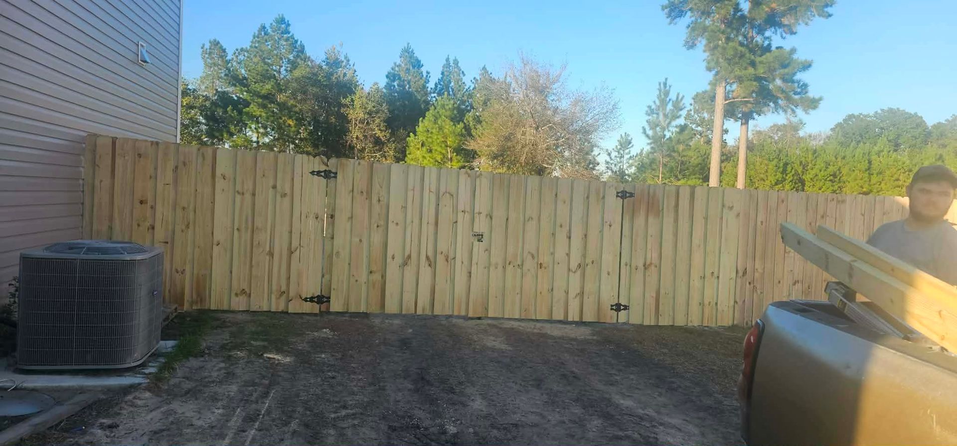 A freshly installed wooden fence runs across a yard next to a house with an air conditioning unit.