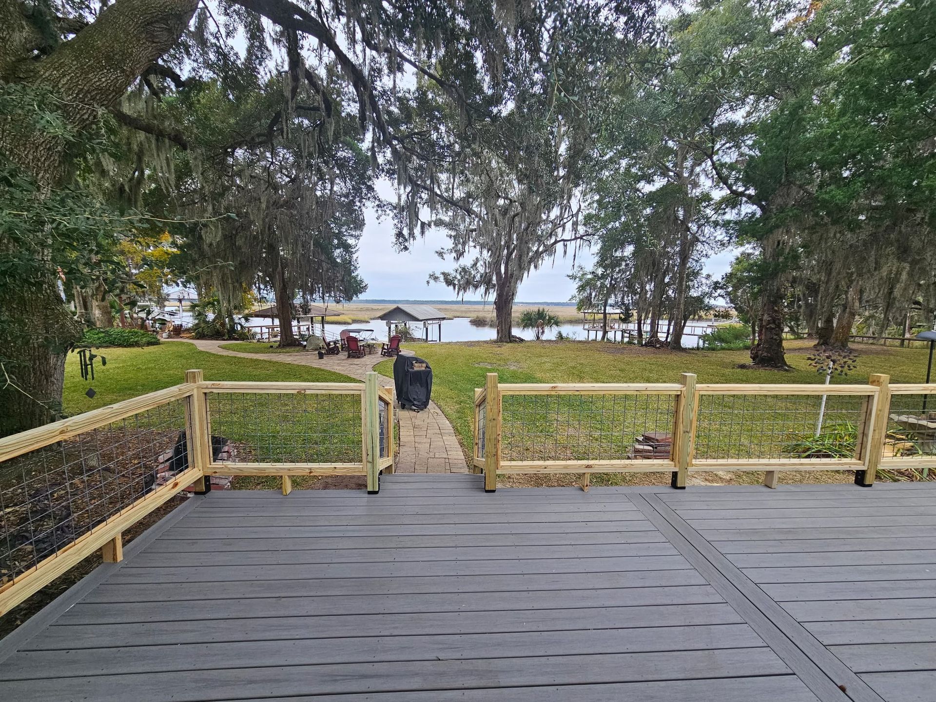 A grey wooden deck with a railing overlooking a grassy lawn leading toward a body of water and distant trees.