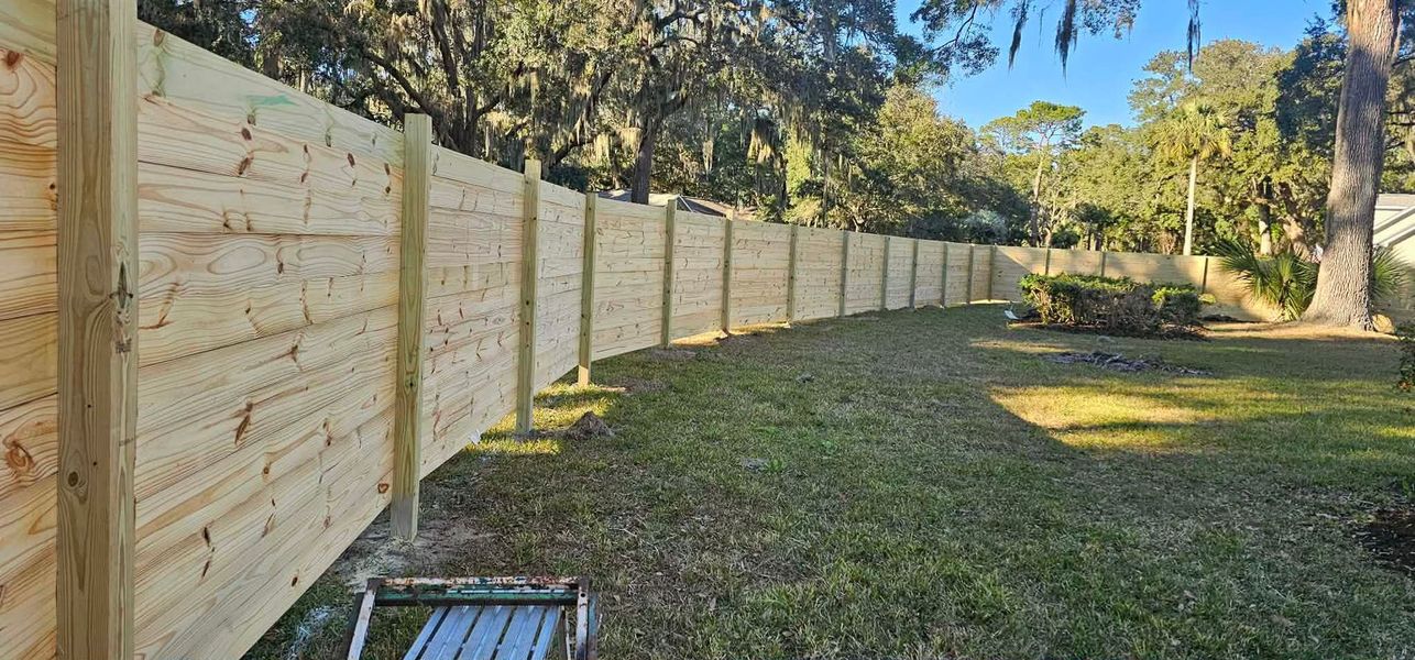A new horizontal-slat wooden privacy fence curves through a sunlit, grassy backyard with trees in the background.