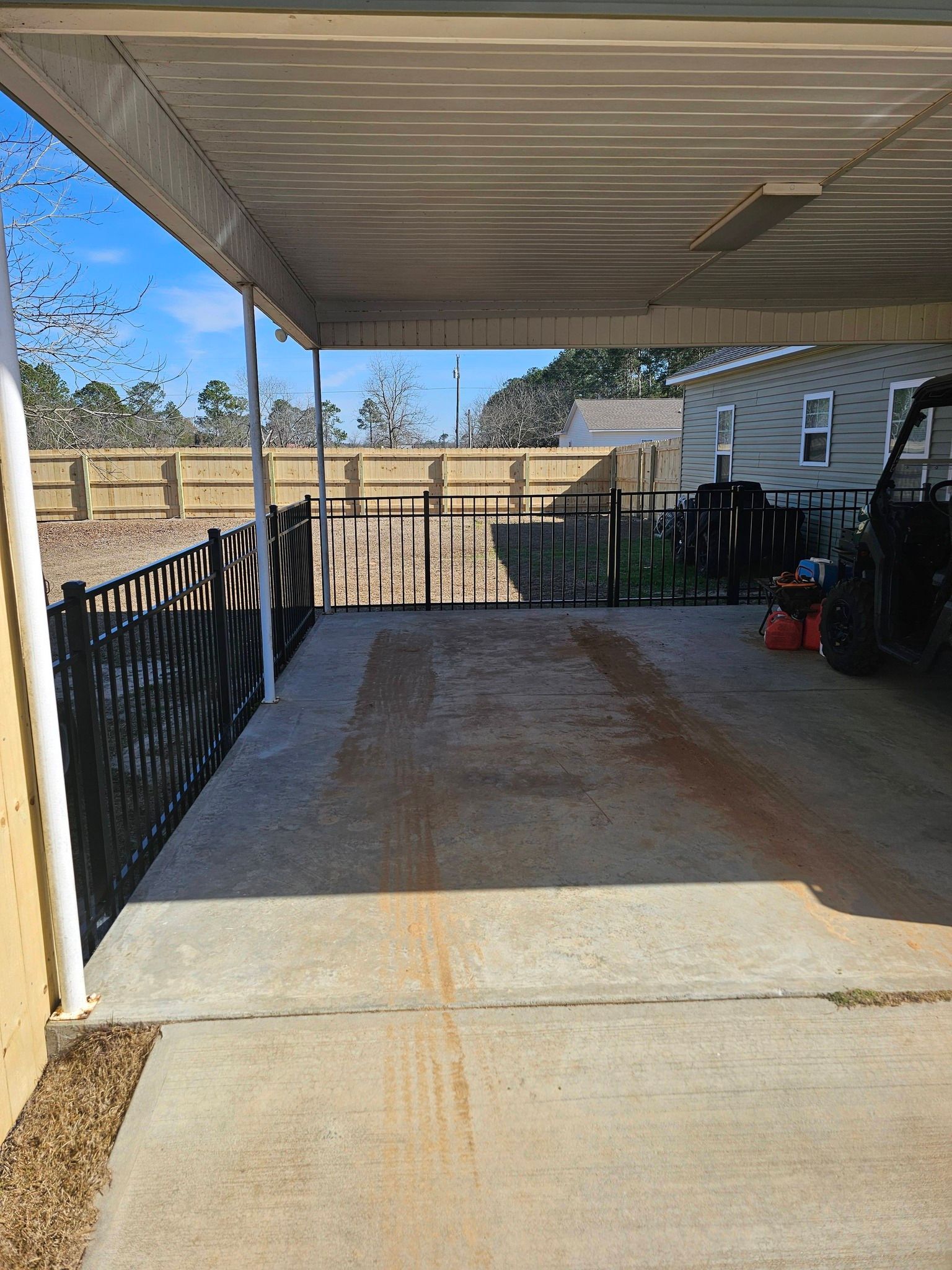 A covered concrete carport with a black metal fence, overlooking an outdoor area with a light wooden fence in the distance.
