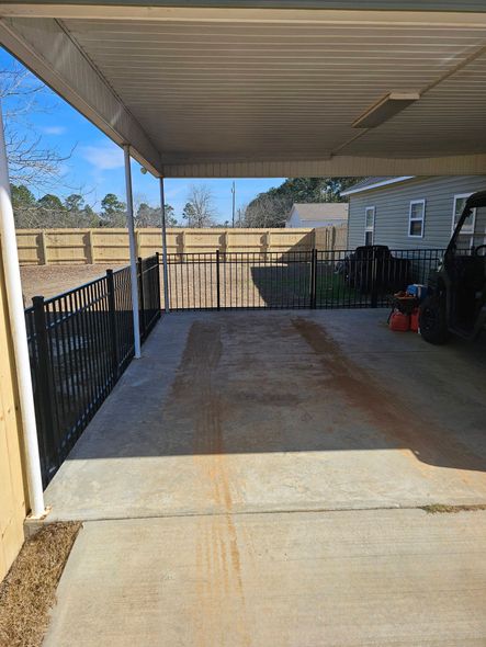 A concrete carport with a black metal fence, leading to a wooden fence and a house in the background.