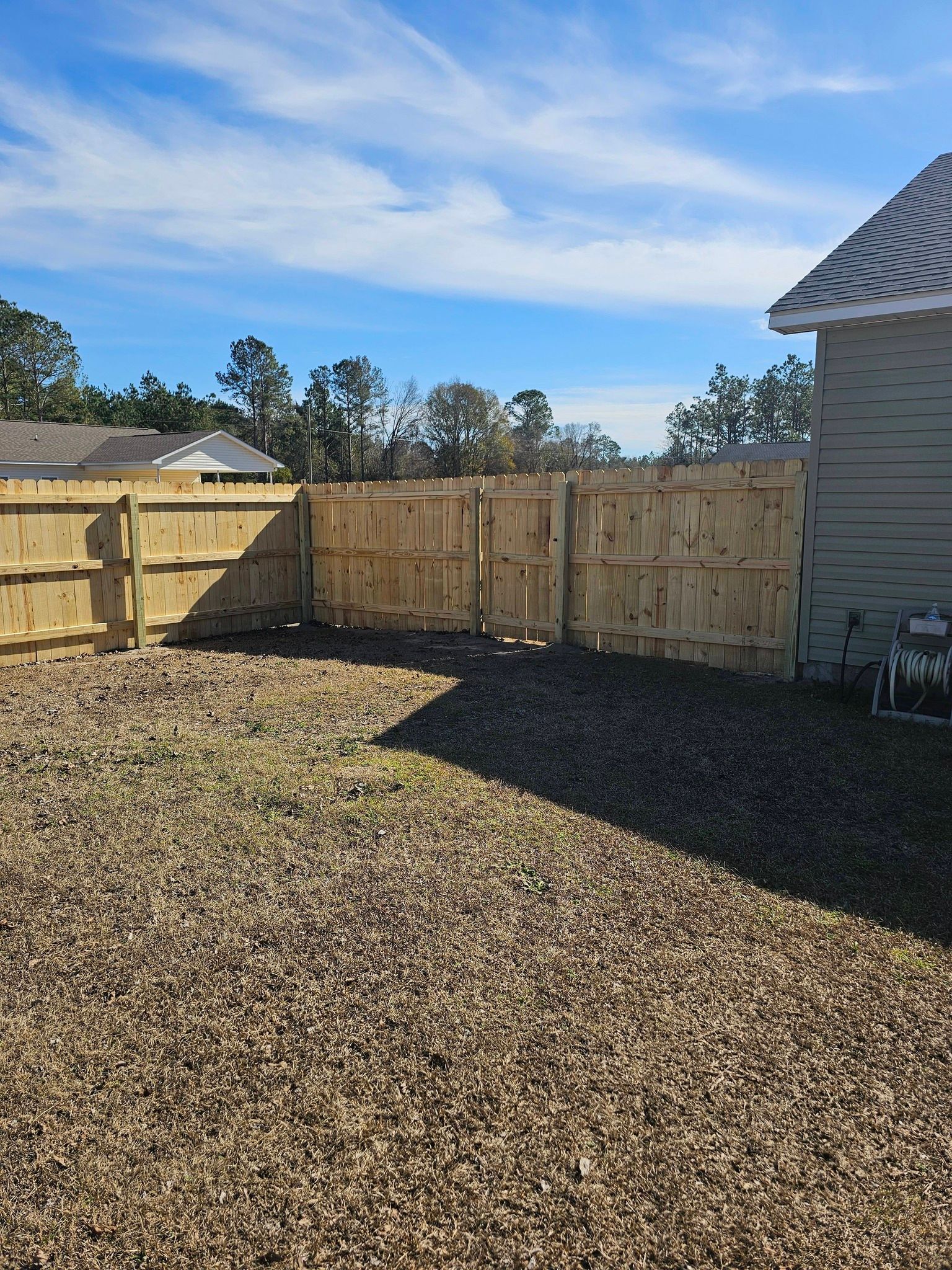 A backyard featuring a gravel ground surface enclosed by a wooden fence, with a house corner visible on the right.
