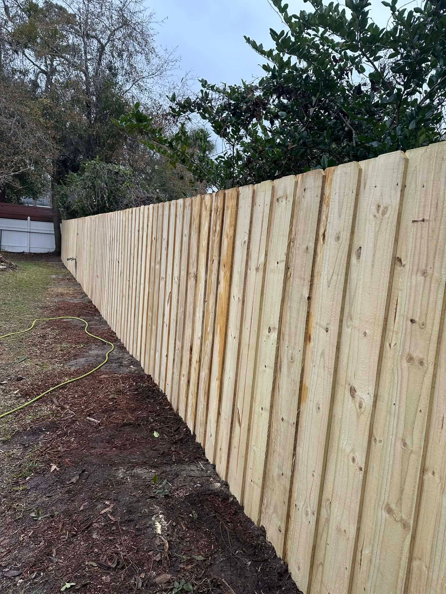 A new light-colored wooden fence runs along a mulched garden bed under a cloudy sky.