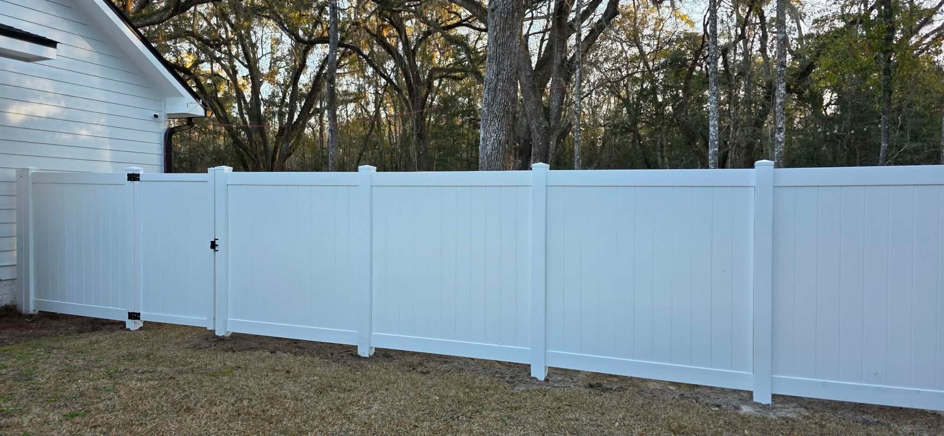 A tall, white vinyl privacy fence stands in a yard in front of a white house and mature trees.