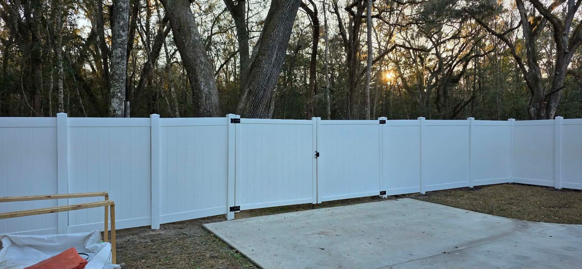 A white vinyl privacy fence with a double gate stands in a backyard bordered by trees at sunset.